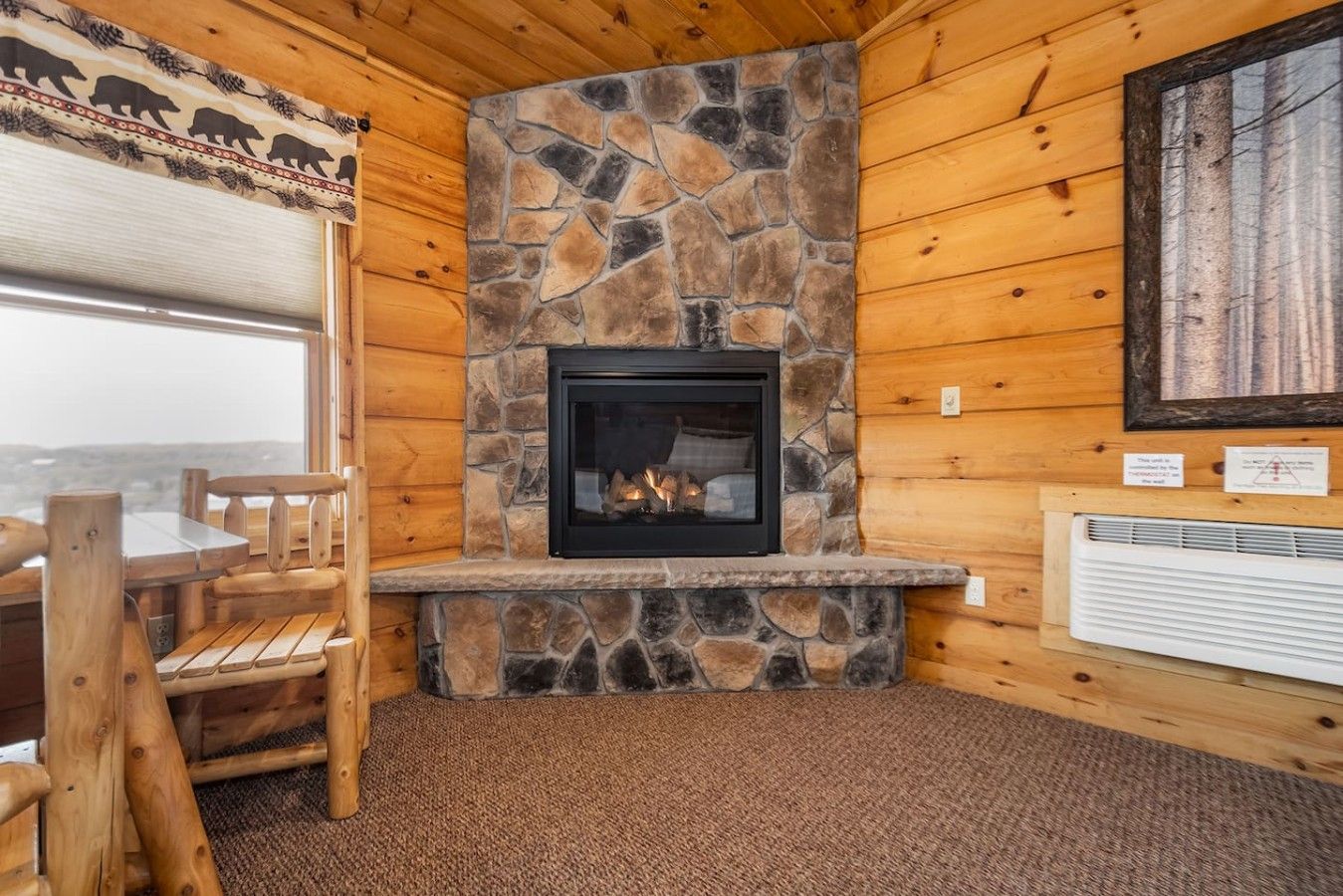 A cozy cabin interior with a stone fireplace, wooden walls, a dining table, and a view through the window.