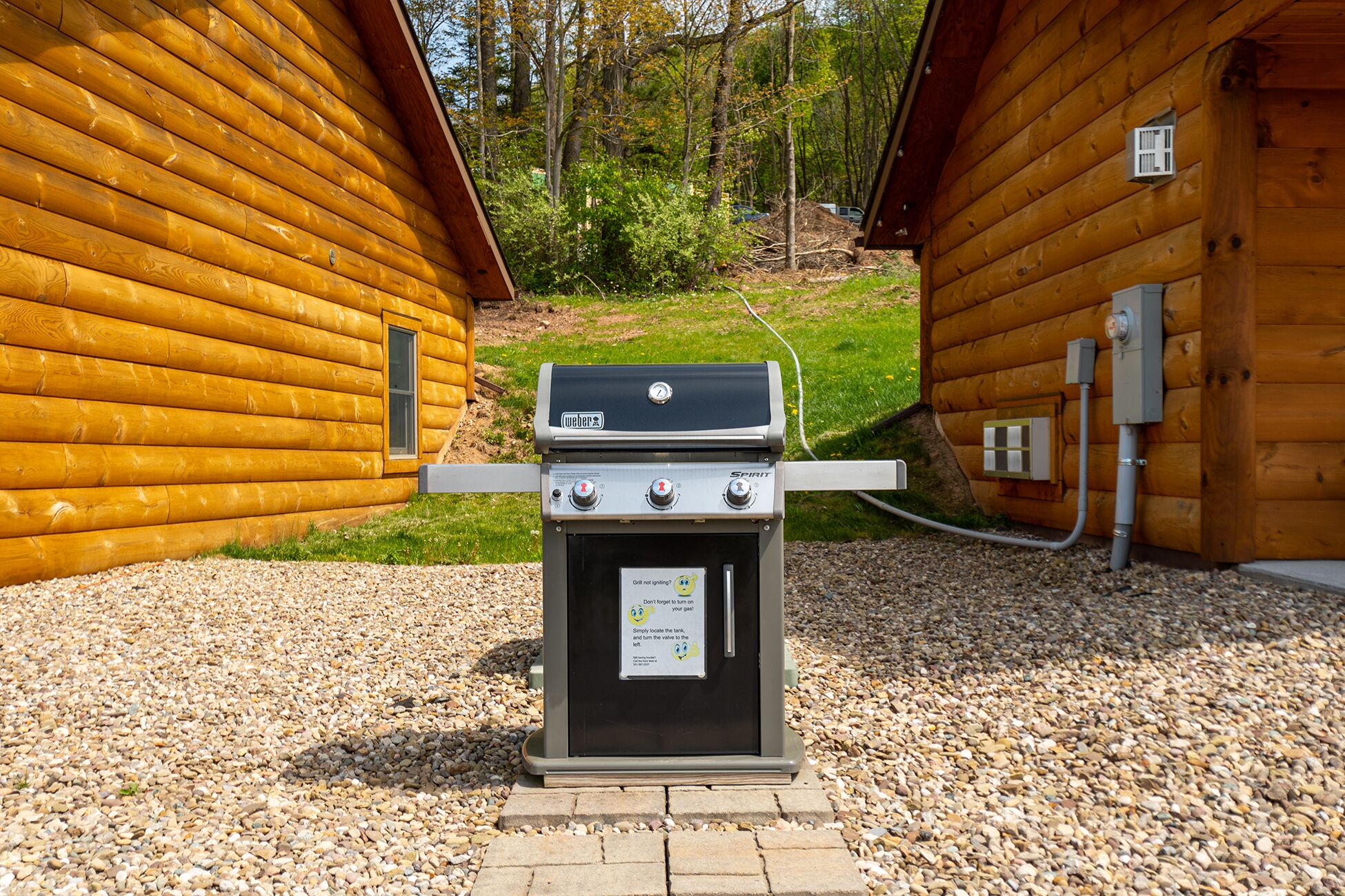 A Weber Spirit grill with a safety warning sign, located between two wooden cabins.