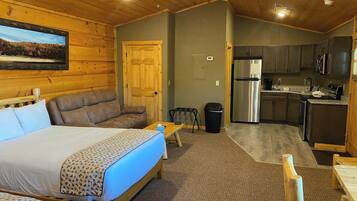 A room with a bed, a sofa, a small table, a trash bin, and a kitchenette area.