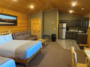 A room with a bed, a sofa, a small table, a trash bin, and a kitchenette area. - Grandview Cabin Suite 29-Full Kitchen (McHenry)