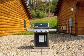 A Weber Spirit gas grill with a built-in oven, positioned on a gravel surface between two wooden cabins. - Grandview Cabin Suite 29-Full Kitchen (McHenry)