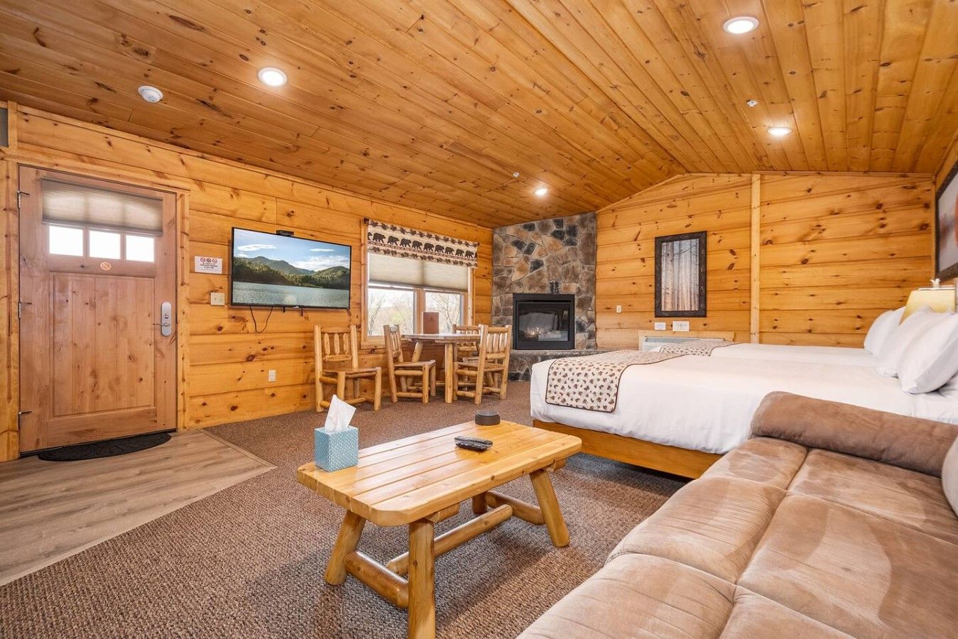 A log cabin interior with a fireplace, a bed, a sofa, a dining table with chairs, and a television mounted on the wall.