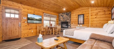 Interior de una cabaña de madera con chimenea, una cama, un sofá, una mesa de comedor con sillas y un televisor en la pared.