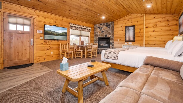 A cozy cabin interior with a wooden ceiling, a stone fireplace, a television, a dining area, and a bed. - Grandview Cabin Suite 29-Full Kitchen (McHenry)