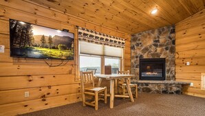 A cozy cabin interior with a stone fireplace, wooden walls, a dining table with chairs, a television mounted on the wall, and a window with a view of the outdoors. - Grandview Cabin Suite 30-Hot Tub, Dog Friendly (McHenry)
