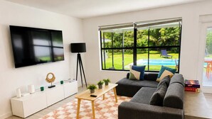 Uma sala de estar moderna com televisão de tela plana, sofá, mesa de centro e uma grande janela com vista para uma piscina.