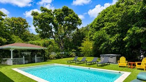 Uma piscina retangular com borda branca, rodeada por grama sintética verde. Em segundo plano, espreguiçadeiras e um gazebo.