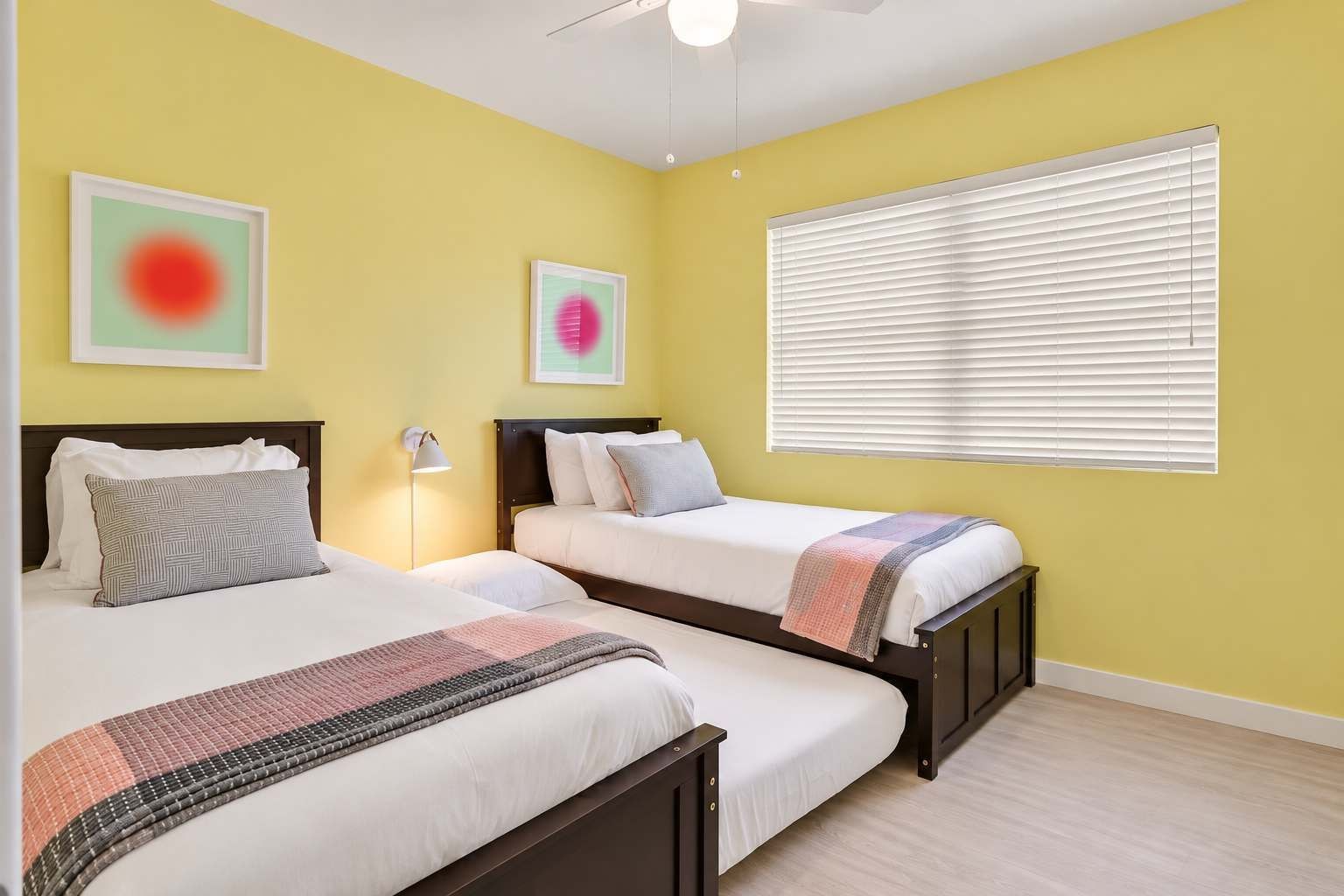 A hotel room with two beds, a ceiling fan, a window with blinds, and artwork on the walls.