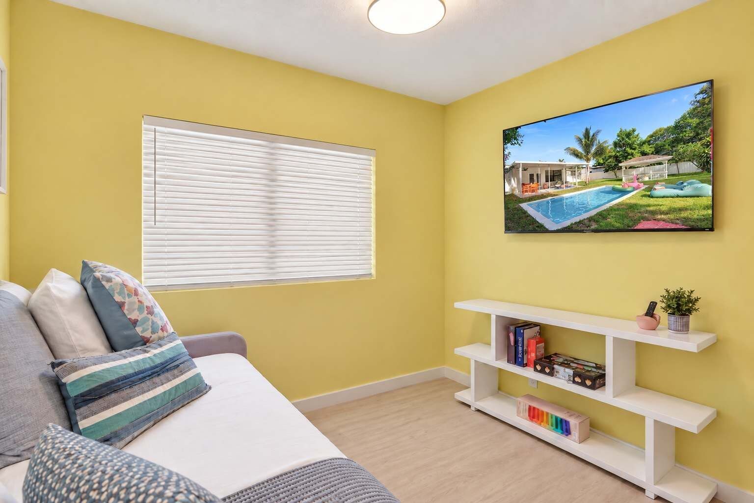 Una habitación con una pared amarilla, un sofá blanco con cojines estampados, un mueble blanco para televisión con libros y una planta, y una ventana con persianas.