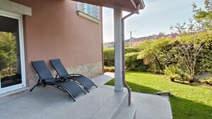 A patio with two lounge chairs, a covered area with a glass door, and a view of a green garden and residential buildings in the background. - Excepcional Chalet en Perbes (Perbes (San Pedro))