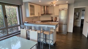 A modern kitchen with a central island, bar stools, a stainless steel refrigerator, and a window overlooking greenery. - Excepcional Chalet en Perbes (Perbes (San Pedro))