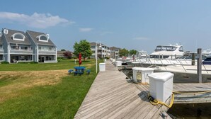 A wooden dock leading to a marina with boats, a grassy area, and a building with multiple windows and balconies. - Waterfront Condo in Spring Lake-Blissful Blue Waterfront (Spring Lake)