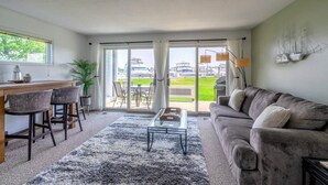 A living room with a grey sofa, a coffee table, a desk with chairs, and a large window overlooking a marina. - Waterfront Condo in Spring Lake-Blissful Blue Waterfront (Spring Lake)