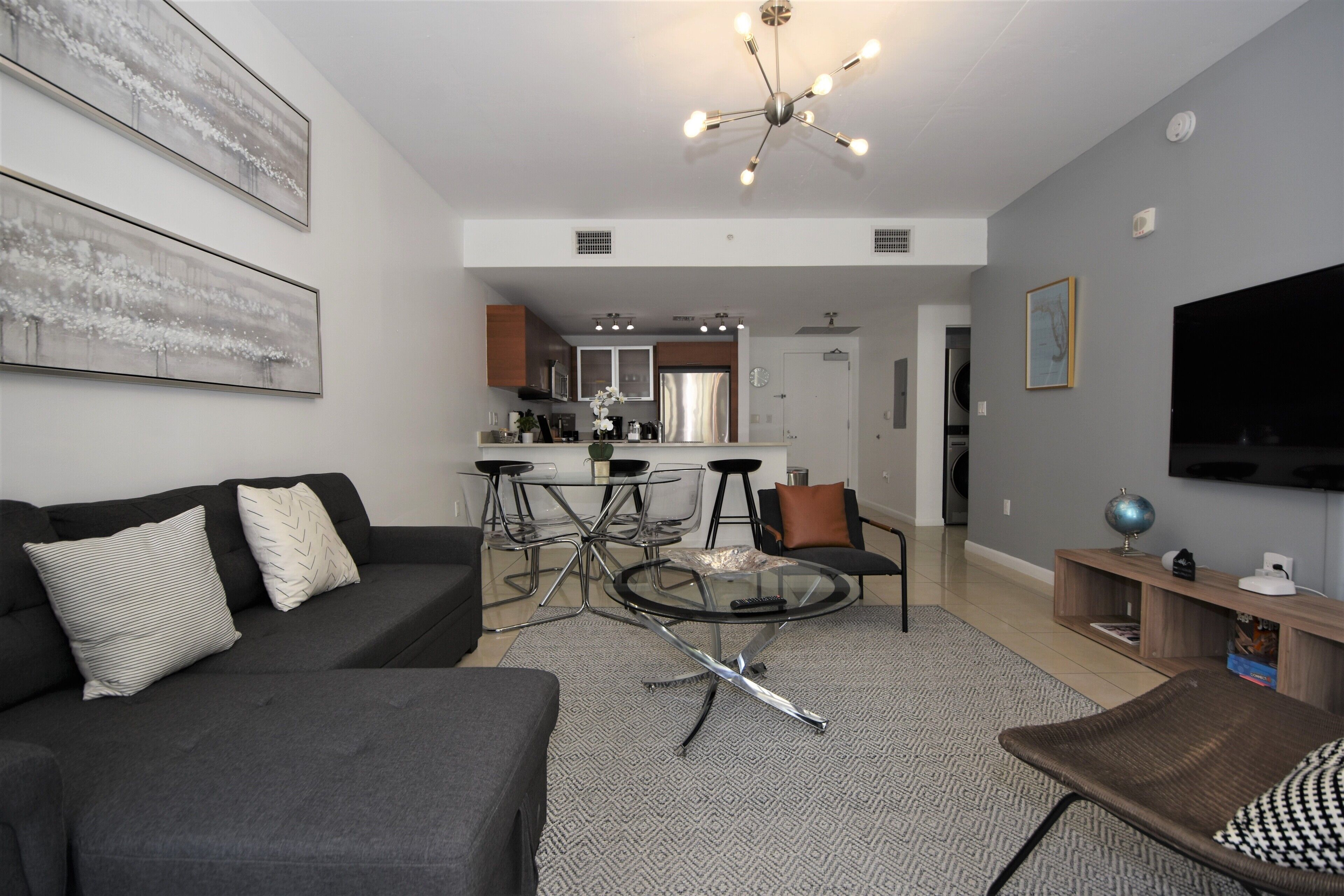 A modern living room with a sofa, a coffee table, and a TV. There is a kitchen in the background with a dining area.