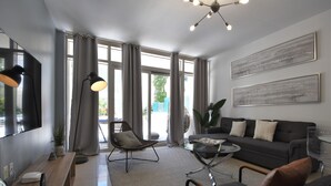 A modern living room with a sofa, coffee table, and a TV. There are large windows with curtains, a ceiling light, and artwork on the wall. - Lovely apt in the heart Midtown (Miami)