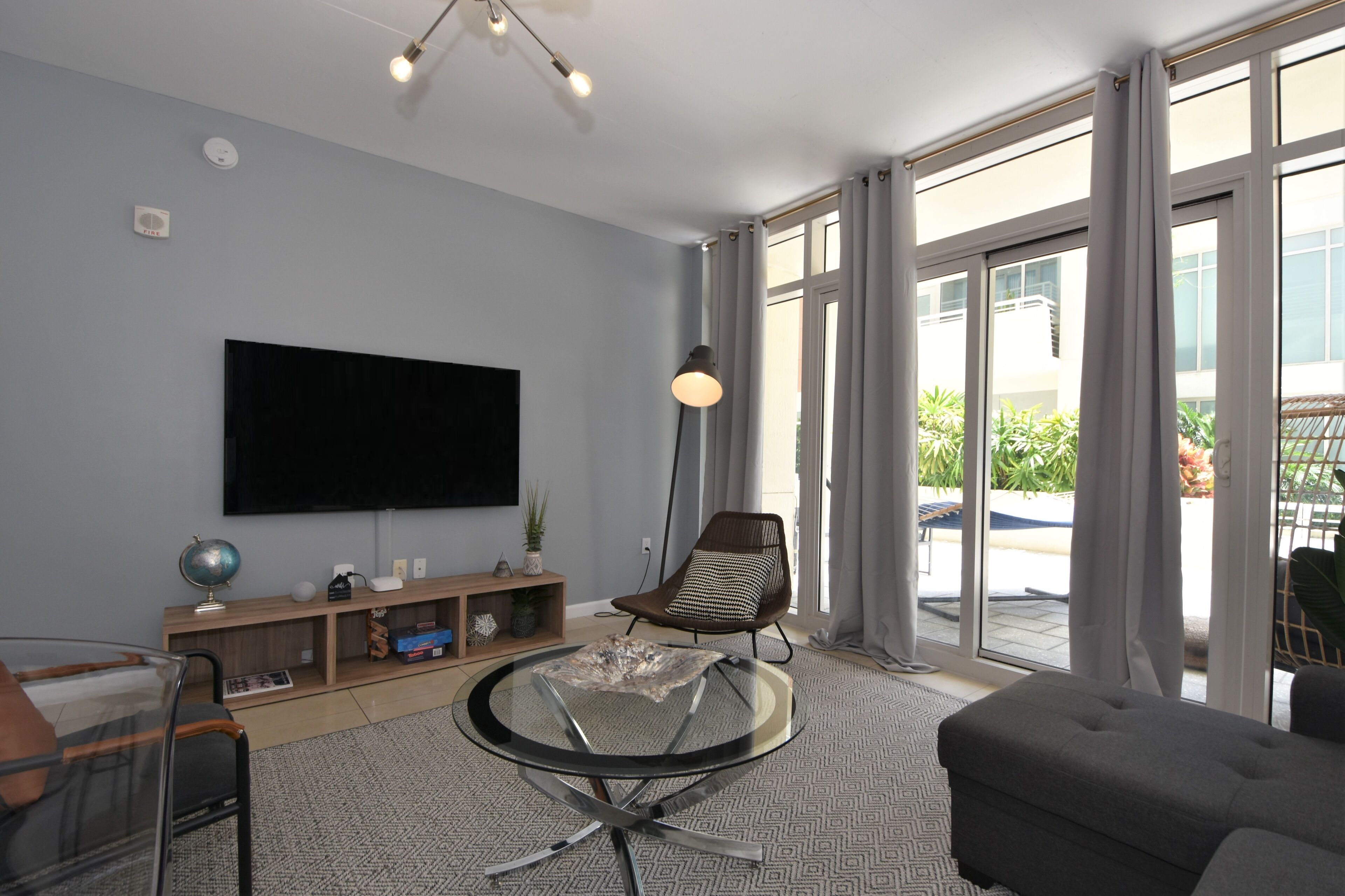 A modern living room with a flat-screen TV, a round glass table, a comfortable sofa, and a large window offering a view of the outdoors.