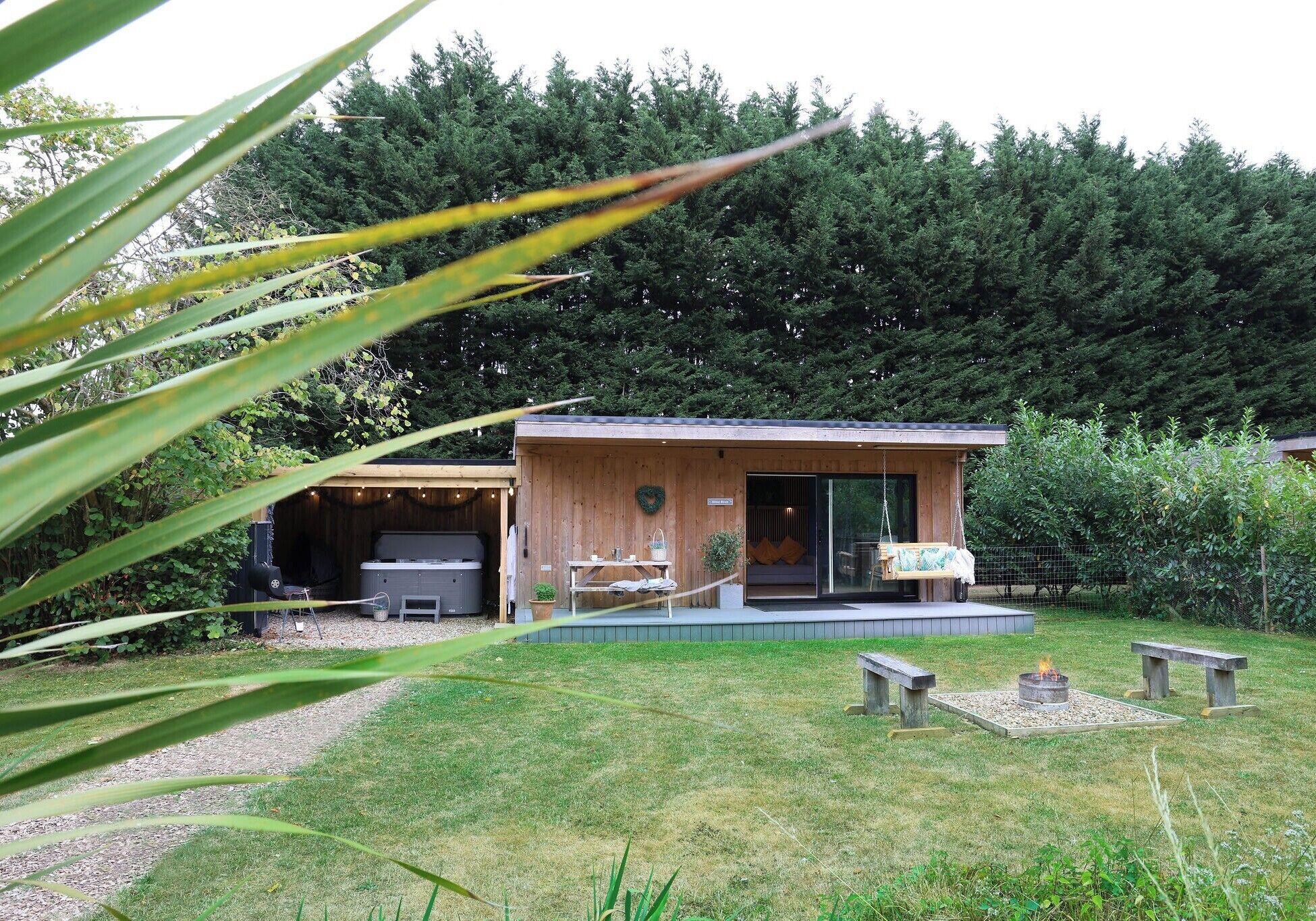 A wooden cabin with a hot tub, a covered outdoor seating area, and a fire pit in the yard.