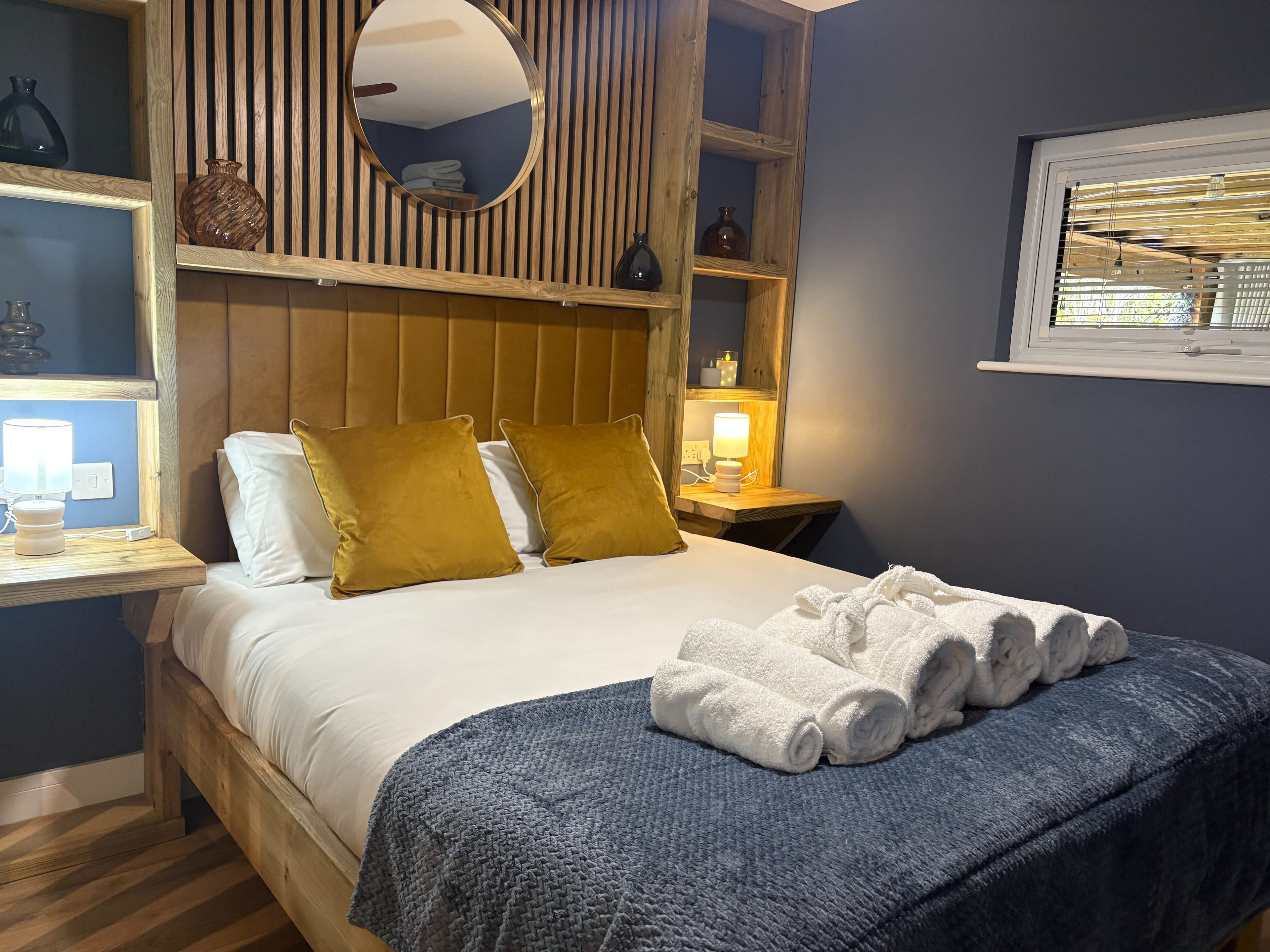 Ein ordentlich bezogenes Bett mit weißen Leinentextilien und zwei senfgelben Kissen, ein Nachttisch mit Lampe und ein Fenster mit Jalousien.