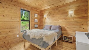 Un dormitorio de cabaña de madera con cama, mesita de noche, lámpara de pared y ventana con vista a follaje.