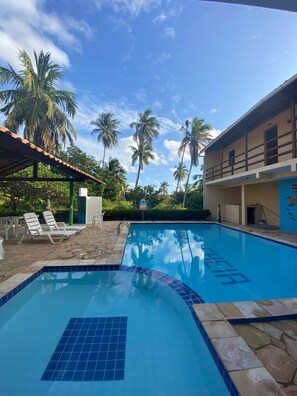 A swimming pool with a tiled deck and lounge chairs, surrounded by palm trees and a building with a tiled roof.