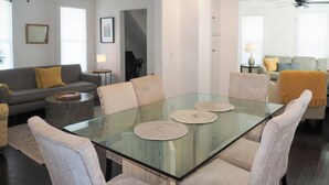 Una sala moderna con una mesa de comedor de vidrio, sillas color beige, un sofá gris con cojines amarillos y una escalera que sube a otro nivel.