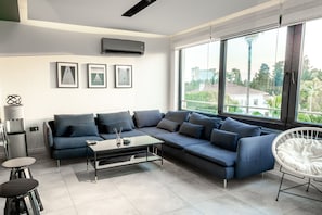 A modern living room with a sectional sofa, a glass coffee table, a white chair, and a large window offering a view of buildings and greenery. - Boho Oldtown Hostel (Antalya)