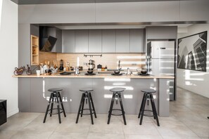 A modern kitchen with grey cabinets, a large island with stools, and a stainless steel refrigerator. - Boho Oldtown Hostel (Antalya)