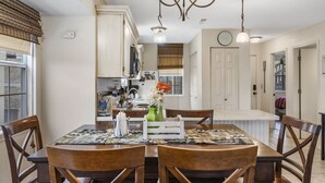 A dining area with a wooden table and chairs, a chandelier, and a kitchen in the background.