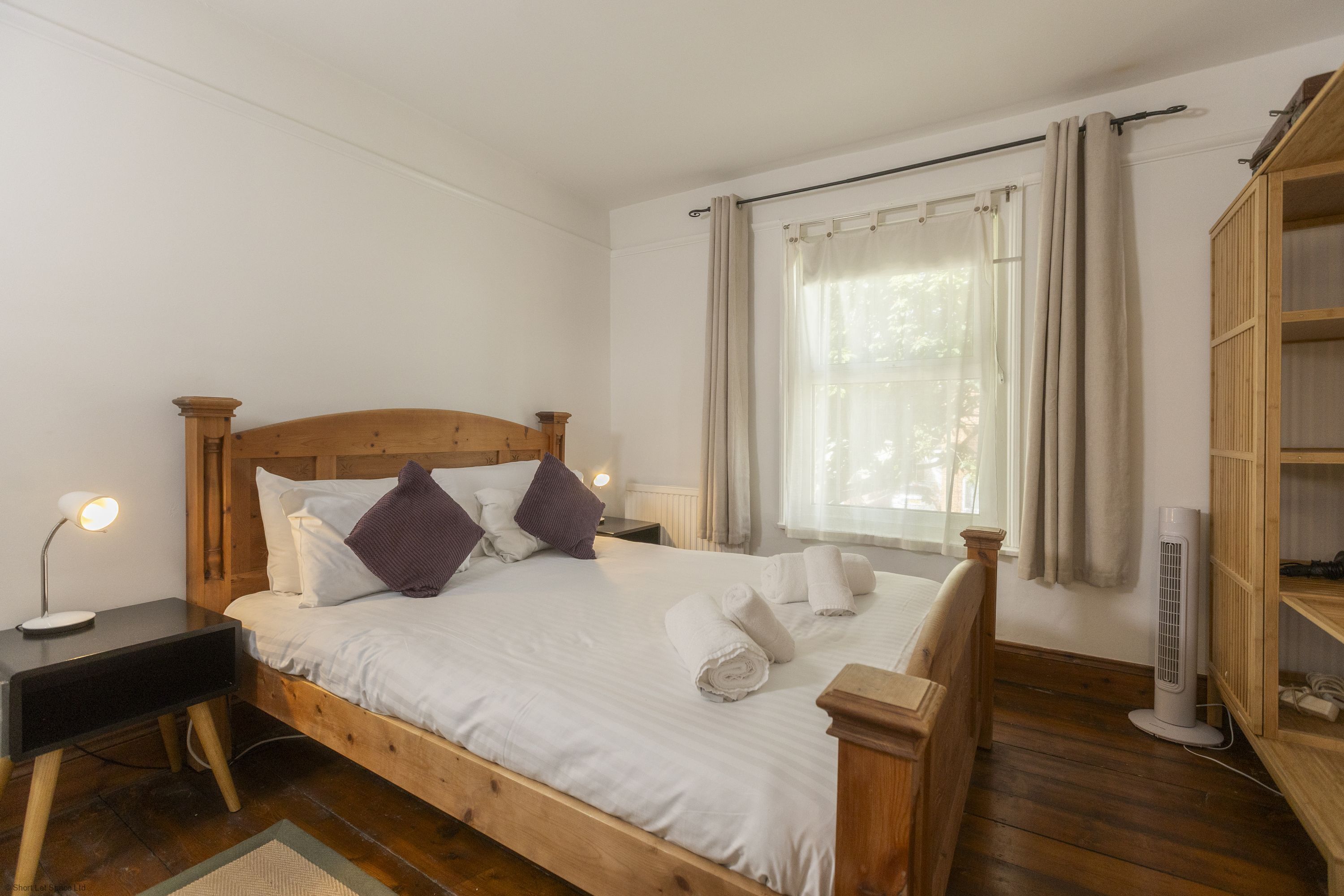 A wooden bed with white bedding, a bedside table with a lamp, a window with curtains, and a wooden wardrobe.