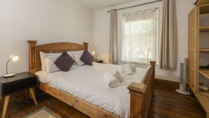 A wooden bed with white bedding, a bedside table with a lamp, a window with curtains, and a wooden wardrobe.