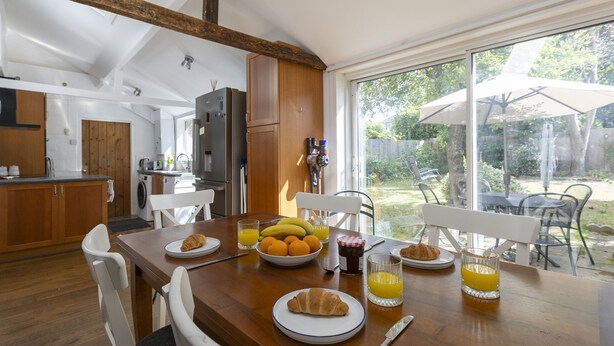 A breakfast table with croissants, oranges, and juice glasses in a bright kitchen with wooden beams and a view of the backyard.