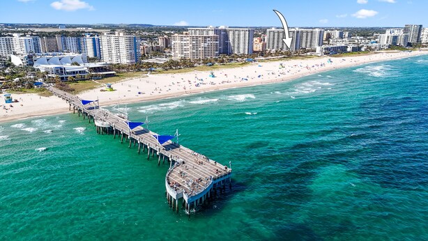 Ein Luftbild von einem Strand mit einem Steg, der sich ins Meer erstreckt, umgeben von Hochhäusern.
