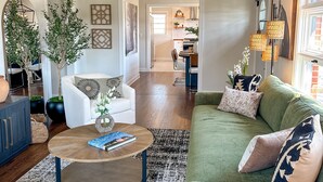 Ein Wohnzimmer mit einem grünen Sofa, einem Couchtisch und einem Deckenventilator. Im Hintergrund ist eine Küche erkennbar.