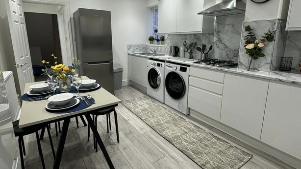 Una cucina moderna con mobili bianchi, un frigorifero in acciaio inox, un forno integrato, un piano cottura a gas e una sveglia appesa al muro.