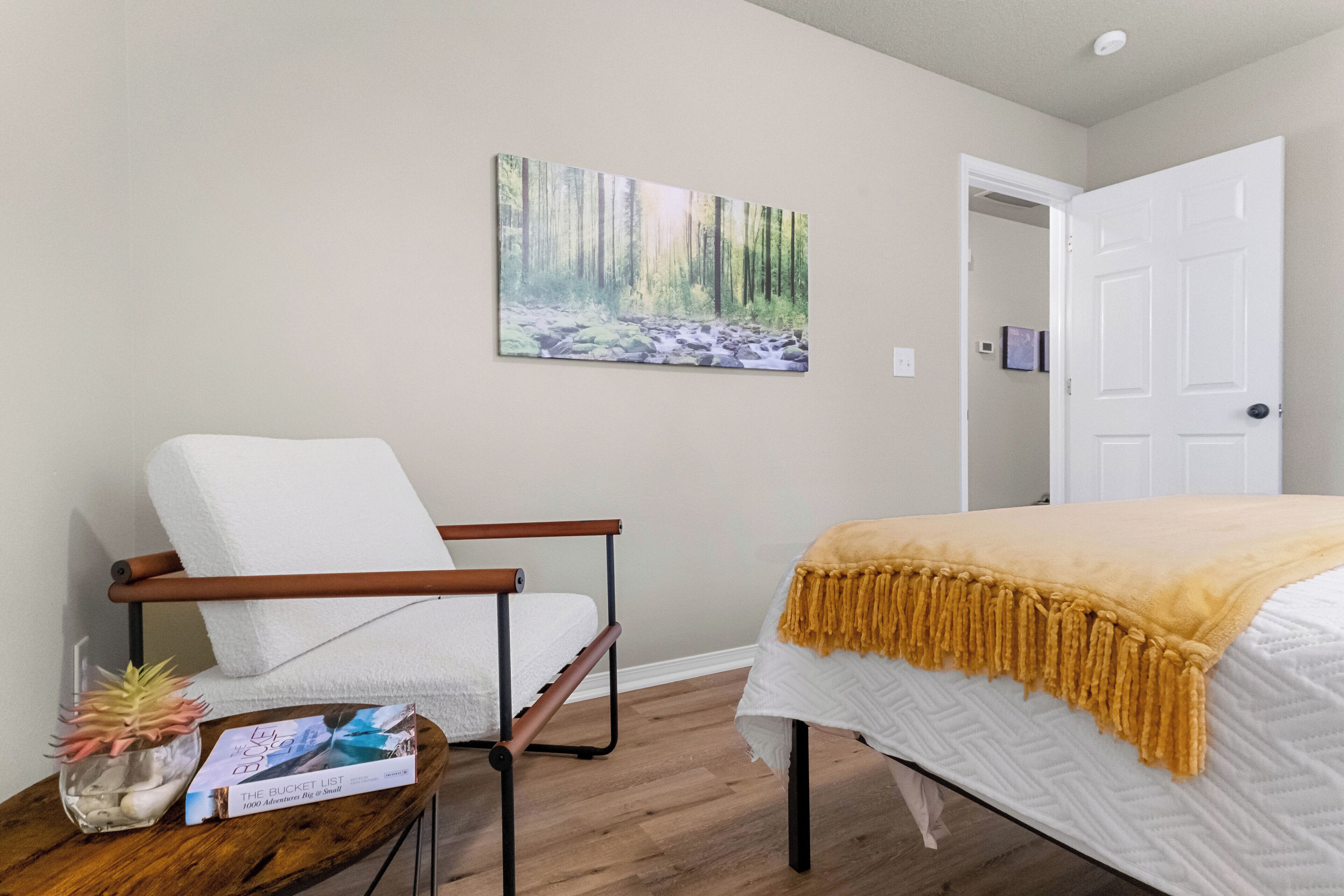 A bedroom with a bed, a white chair, a wooden side table with a plant and a bucket list book, and a painting of a forest on the wall.