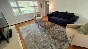 A living room with a large window, a dark blue sofa, a white armchair, a coffee table with a compass design, and a lamp. - Cape Cod Fun - Entire home 3BR up to 9 guests  (Barnstable)