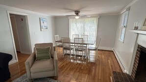 A living room with a beige sofa, a dining table with chairs, a ceiling fan, and a fireplace. - Cape Cod Fun - Entire home 3BR up to 9 guests  (Barnstable)