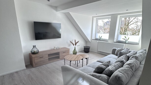 A modern living room with a large grey sofa, a flat-screen TV mounted on the wall, a wooden cabinet, and a small table with a vase. - Ferienwohnung in Obernkirchen (Obernkirchen)