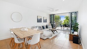 A modern living room with a wooden dining table, white chairs, a grey sofa, and a large sliding glass door leading to an outdoor area.
