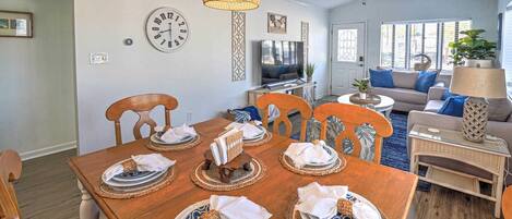 A dining area with a wooden table set for six, a television in the background, and a living area with a sofa and a lamp.