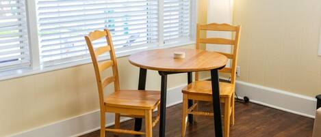 Una habitación con una mesa redonda de madera y dos sillas de madera, una ventana con persianas y una lámpara fijada a la pared.