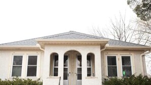 Une maison à deux étages, dotée d’un toit à pignon, d’une entrée centrale avec une double porte et d’un petit porche agrémenté d’une balustrade.