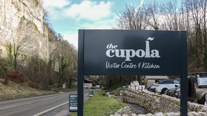 Placa para 'The Cupona Visitor Centre & Kitchen', com um muro de pedra e veículos ao fundo.