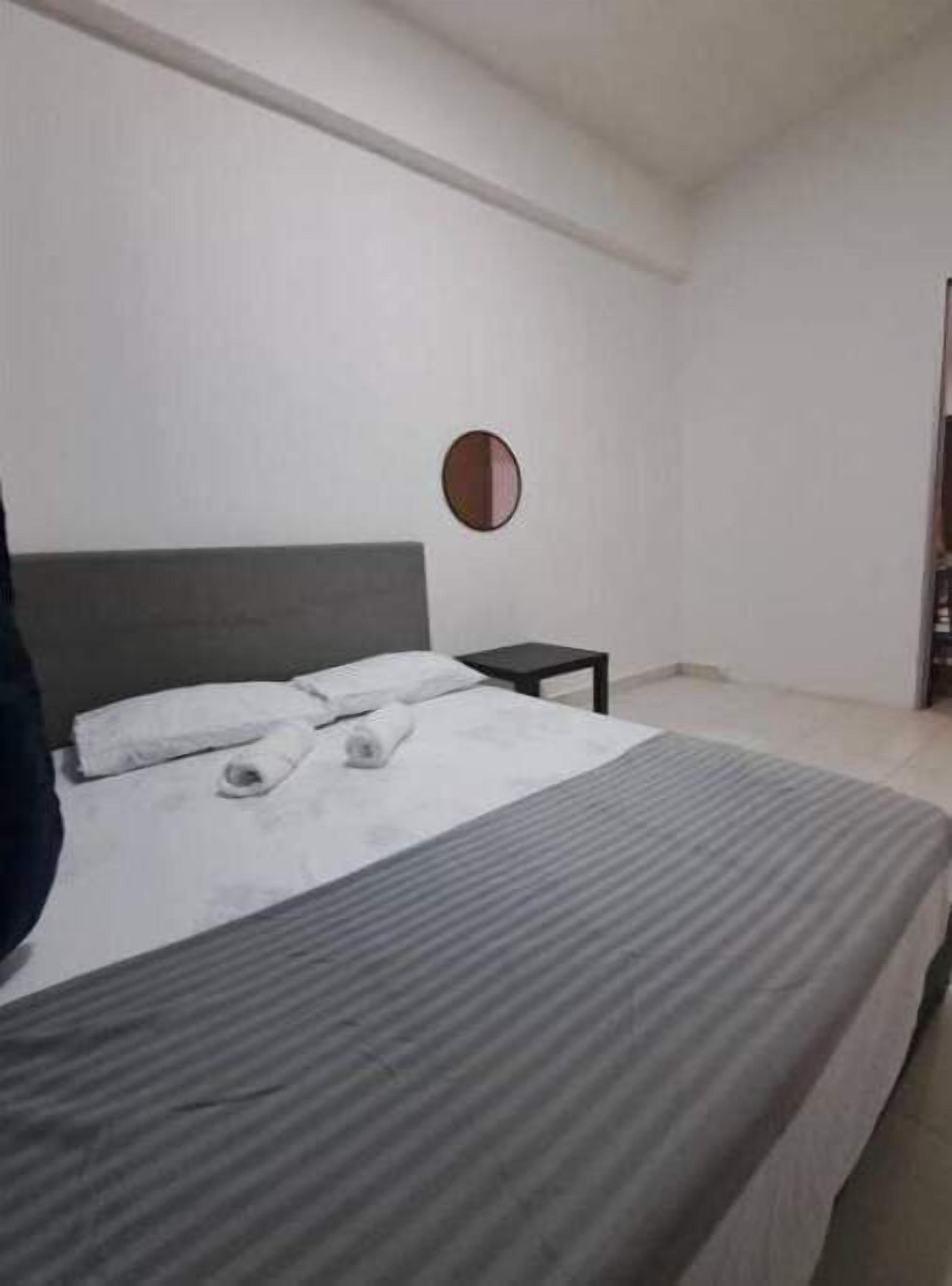 Un lit simple avec une couvre-lit à rayures grises, un oreiller blanc et une serviette enroulée posée sur le lit. Un petit miroir rond est accroché au mur, au-dessus du lit.