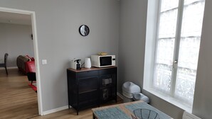 Ein modernes Wohnzimmer mit Holzboden, einem Esstisch mit gewebter Tischmatte, einem schwarzen Metallkorb, einer Mikrowelle und einem kleinen weißen Gerät auf einer Holzkommode.