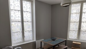 Ein Zimmer mit Tisch, Bank und einem kleinen Hocker. Zwei Mülleimer und eine Heizung sind vorhanden.
