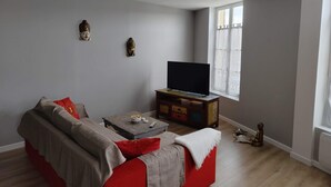 Ein Wohnzimmer mit Sofa, Couchtisch, Fernseher auf Holzfuß und einem Fenster mit Vorhängen.