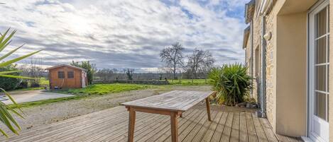A wooden deck with a table, a building with a window, and a grassy area in the background.