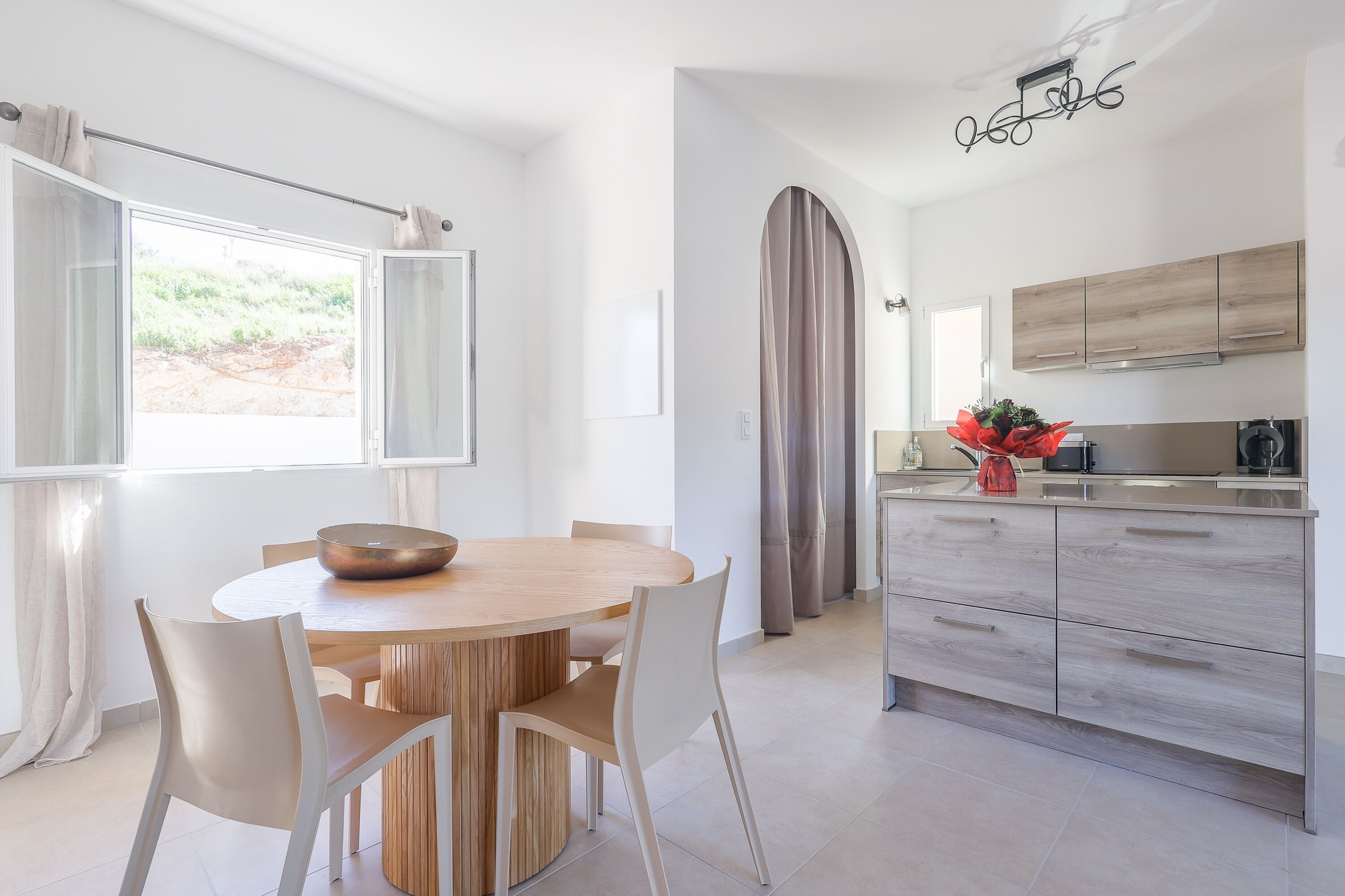 A modern kitchen with wooden cabinets, a dining area with a round table and chairs, and a large window with curtains.