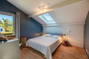 Ett sovrum med ett takfönster, en säng, ett sängbord, en lampa och ett fönster med utsikt över palmer.
